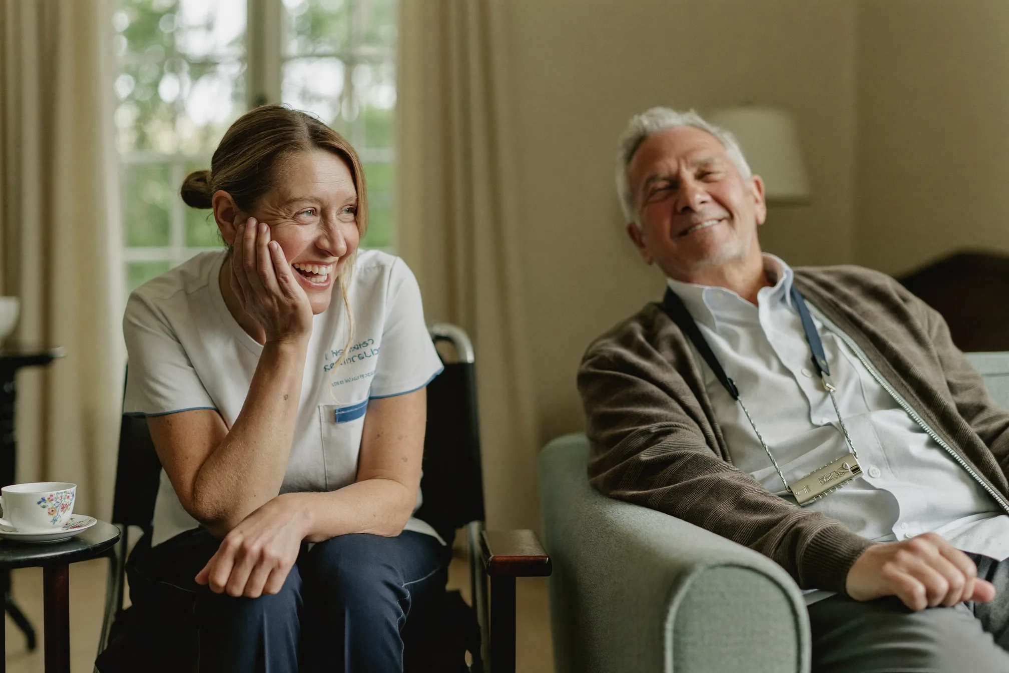 A support worker and an elderly man laughing together over tea in a cosy living room
