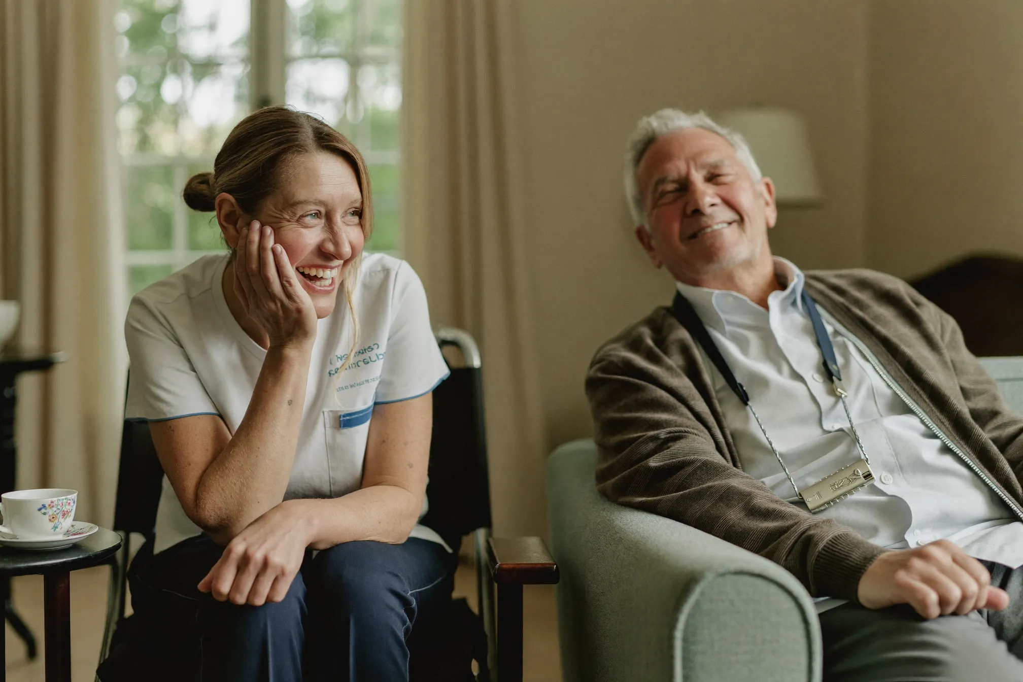 A support worker and an elderly man laughing together over tea in a cosy living room