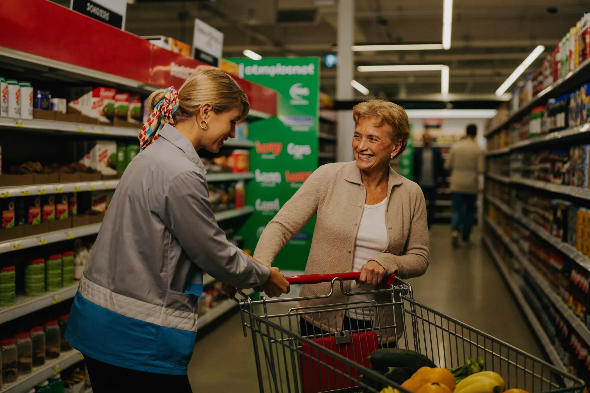 A support worker helping an elderly woman with a shopping trolley in a supermarket aisle