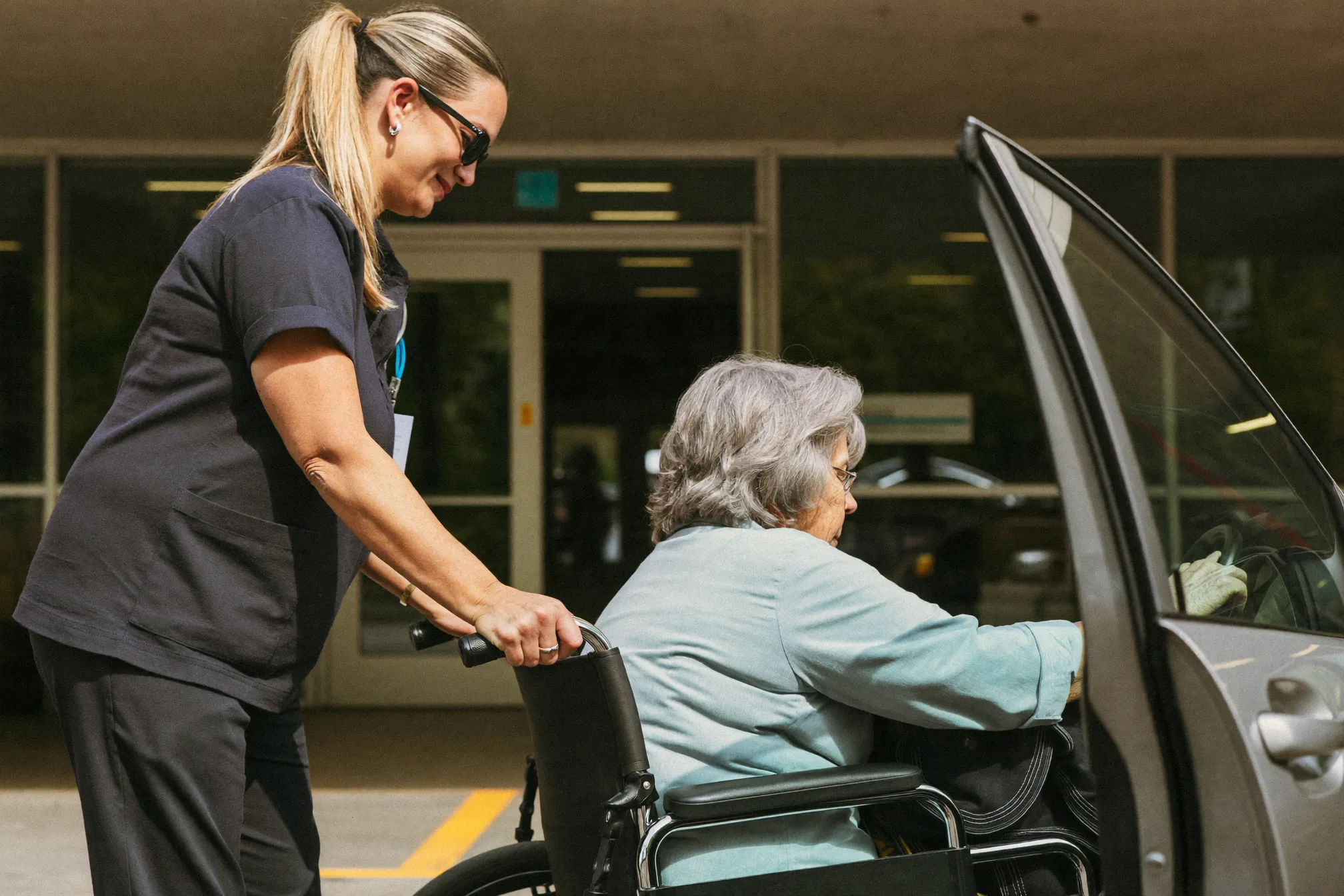 A support worker helping an elderly woman from a wheelchair into a car outside a hospital